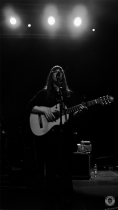 Sängerin mit Gitarre am Mikrofon. Frontal fotografiert in schwarz weiß.