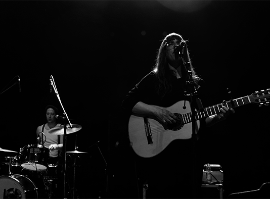 Sängerin mit Gitarre am Mikrofon. Frontal fotografiert in schwarz weiß.